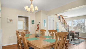 A dining area with a wooden table and chairs, a chandelier, and a staircase leading to another level. - Shell Cottage | East Side Assateague Channel Views w/ Decks, Outdoor Shower, Boat Parking & Pets (Chincoteague Island)