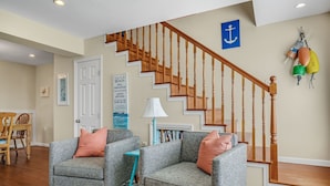A living room with two grey armchairs, a small blue side table, a wooden staircase, and a blue anchor decoration on the wall. - Shell Cottage | East Side Assateague Channel Views w/ Decks, Outdoor Shower, Boat Parking & Pets (Chincoteague Island)