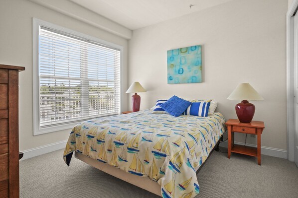 A bedroom with a large bed, two bedside tables with lamps, a window with blinds, and a wall painting.