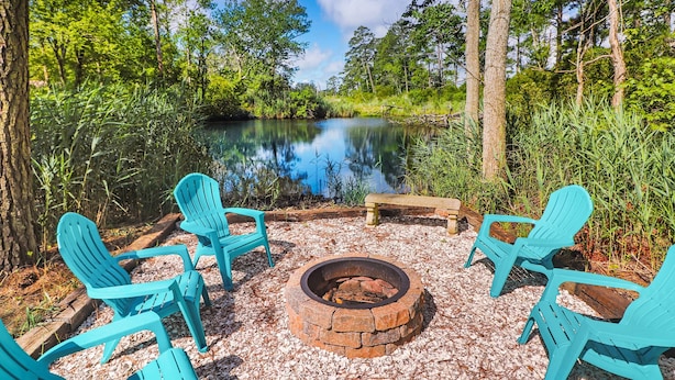 Un espace extérieur paisible, aménagé avec un foyer et des fauteuils Adirondack, entouré d’un paysage naturel et bordé d’une étendue d’eau calme.