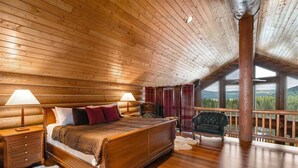 A log cabin bedroom with a wooden bed, nightstands, a chair, and a balcony with a view.