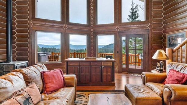 A log cabin interior with a fireplace, leather furniture, a wooden dining table, and large windows offering mountain views.