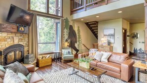 A spacious living room with a stone fireplace, a large window with a view of trees, a leather sofa, and a wooden staircase. - Gold Baron Lodge (Lead)