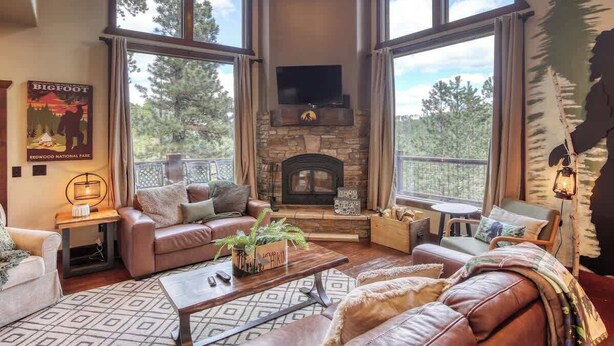 A spacious living room with a stone fireplace, leather sofas, and large windows offering a view of the outdoors. - Gold Baron Lodge (Lead)