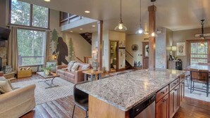 A spacious living area with a large island, wooden flooring, and a view of the outdoors through large windows. - Gold Baron Lodge (Lead)