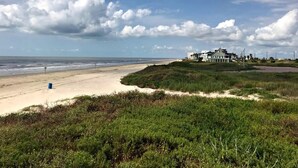 A beach with a large house on a grassy hill, a sandy beach, and the ocean in the background. - Lollygaggin by the Beach! Fenced Yard, Outdoor Bar! (Galveston)