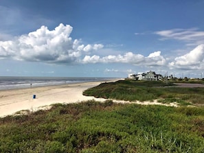 A beach with a large house on a grassy hill, a sandy beach, and the ocean in the background. - Lollygaggin by the Beach! Fenced Yard, Outdoor Bar! (Galveston)