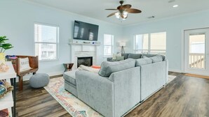 A living room with a gray sofa, a fireplace, a ceiling fan, and a television. - Lollygaggin by the Beach! Fenced Yard, Outdoor Bar! (Galveston)