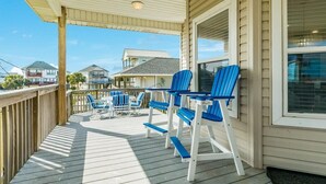 A covered deck with blue chairs and a table, wooden railings, and a view of neighboring houses. - Lollygaggin by the Beach! Fenced Yard, Outdoor Bar! (Galveston)