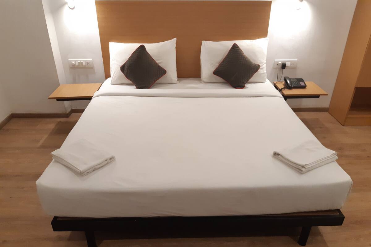 Ein ordentlich bezogenes Bett mit weißen Leinen und zwei dunklen Kissen, ein Nachttisch mit Telefon und ein Beistelltisch mit Lampe.