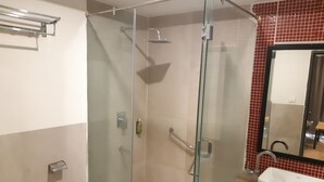 A glass-enclosed shower with a handrail, a white sink, and a mirror with a red-tiled backsplash.