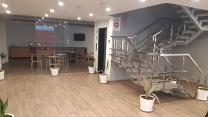 A modern hotel lobby with wooden flooring, potted plants, a staircase, and a conference room visible through a glass partition.