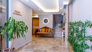 Una hall d'albergo moderna con pavimento in marmo, una comoda poltrona in legno con cuscini colorati, una pianta in vaso e una scalinata che conduce ai piani superiori.