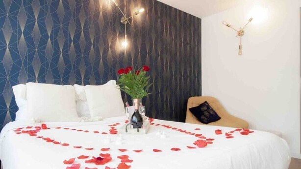 A neatly made bed with white bedding and red rose petals, a vase with red roses, and a geometric patterned wall.