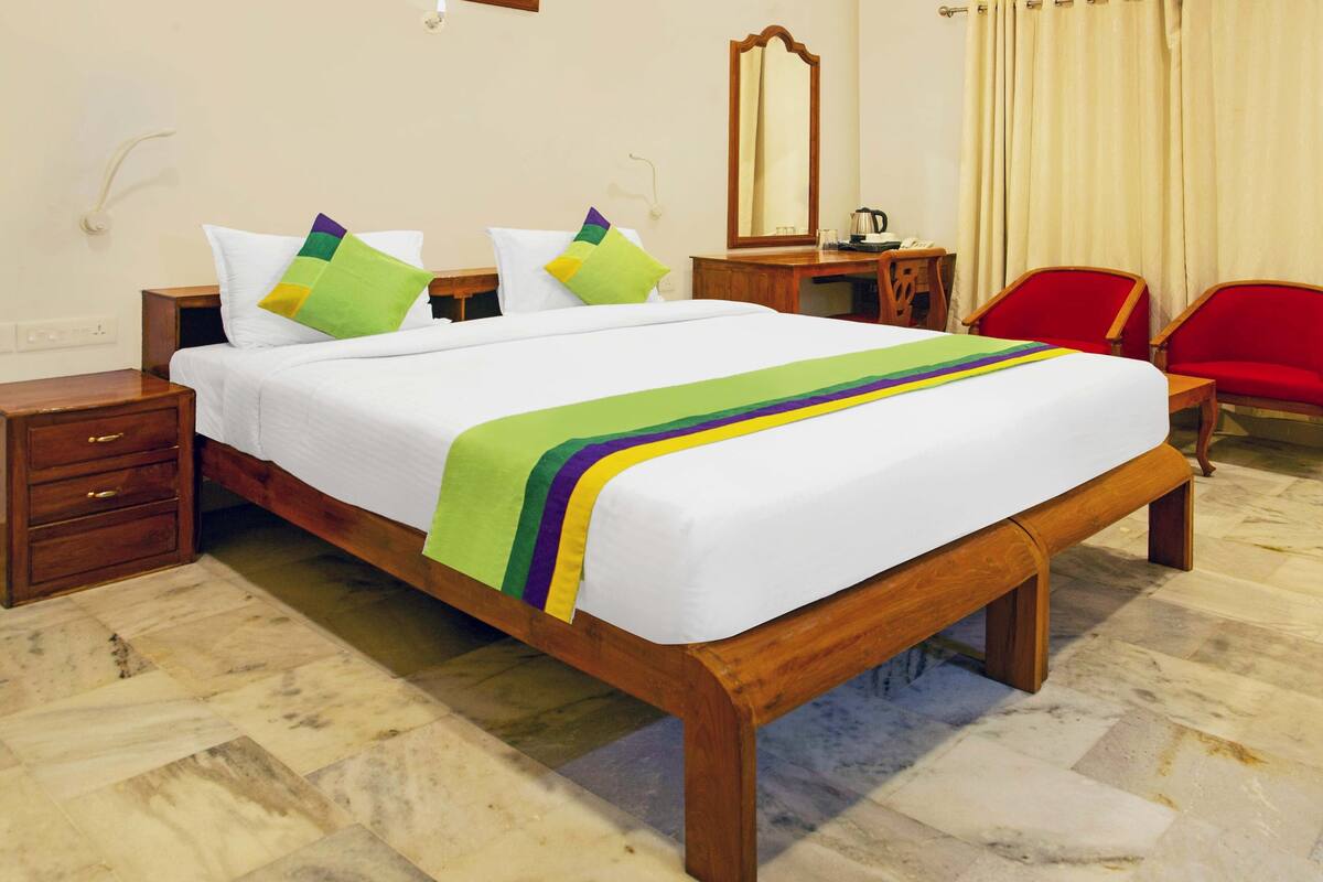 Una cama bien hecha con un edredón colorido, una mesita de noche de madera, un escritorio de madera con una silla, y una silla roja.
