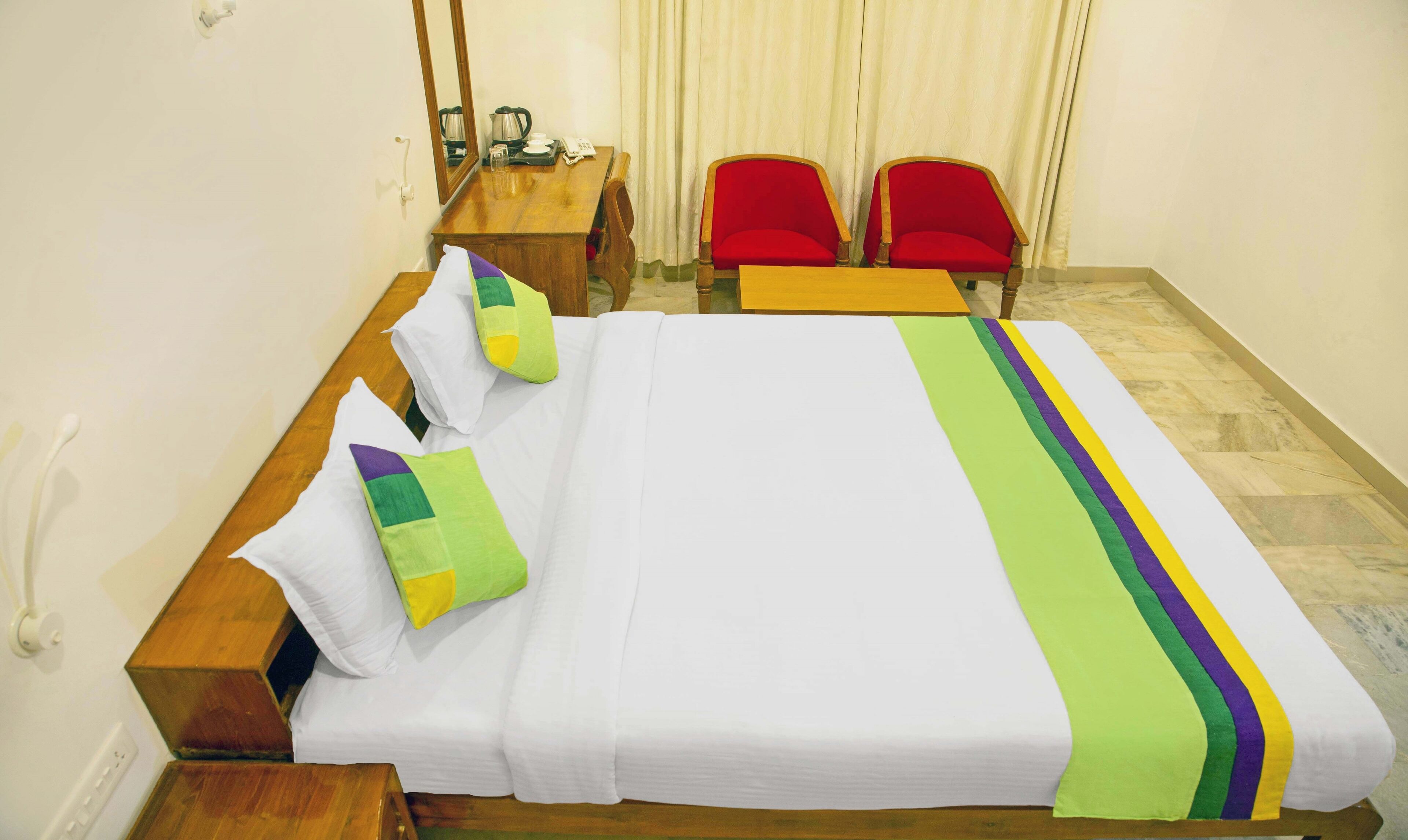 A hotel room with a bed, two red chairs, a wooden table, a mirror, and a telephone.