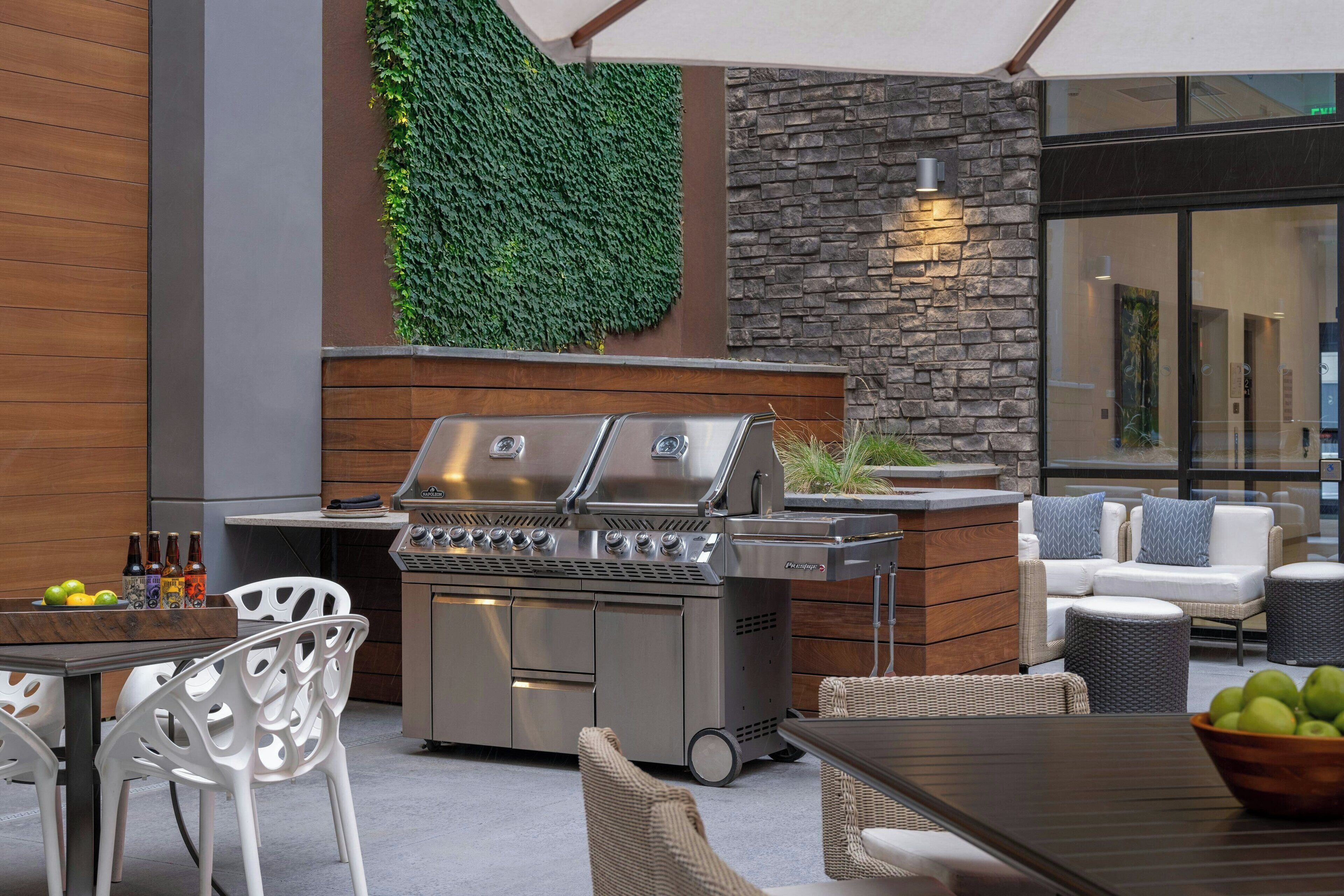 Un patio al aire libre con una parrilla de acero inoxidable, mobiliario de mimbre y una pared de piedra con vegetación.