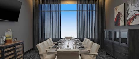 A conference room with a long table, leather chairs, a chandelier, a TV, and large windows with curtains.