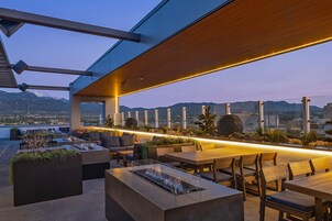 An outdoor seating area with a fire feature, wooden tables, and chairs, overlooking a cityscape and mountains.