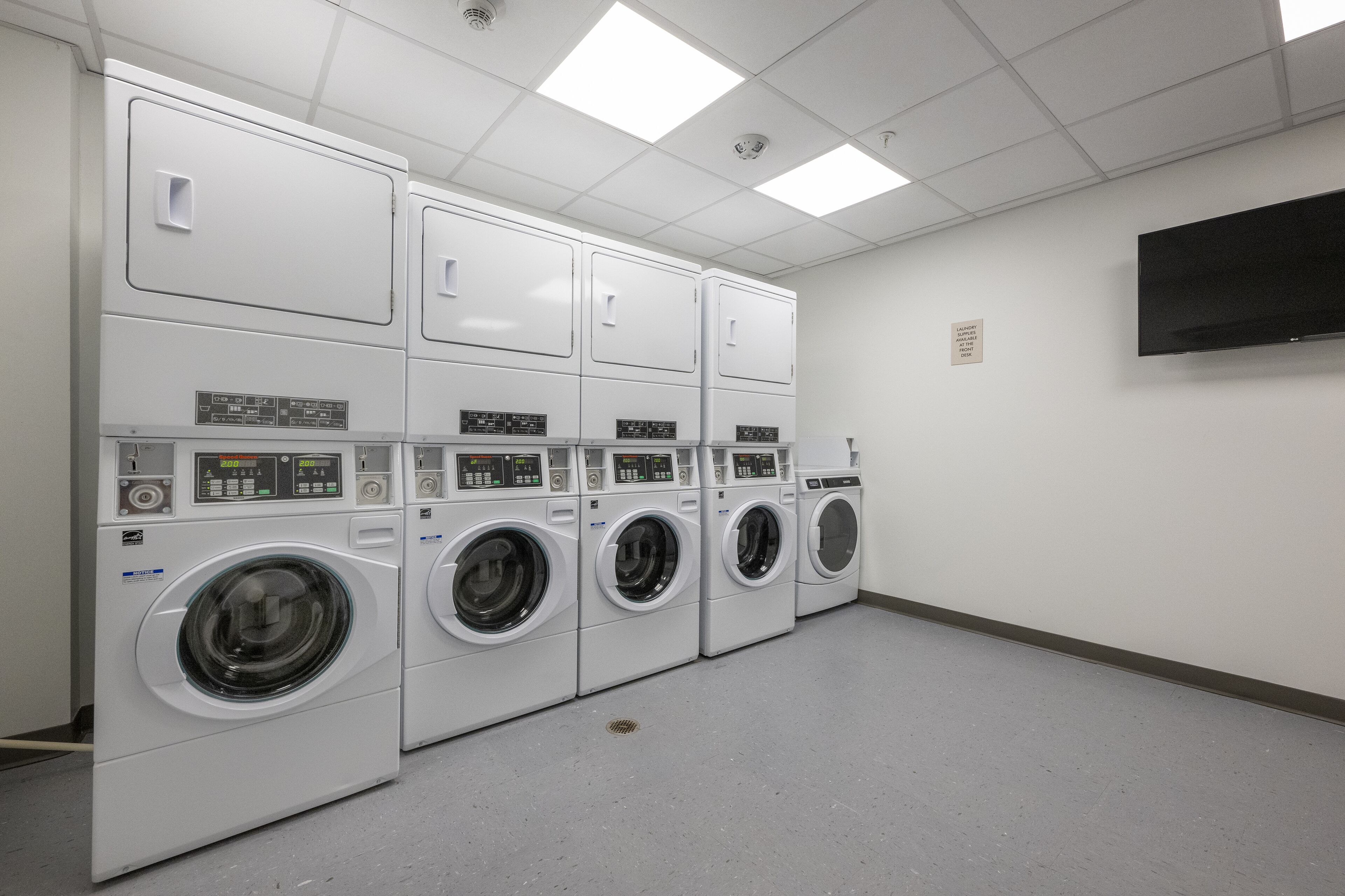 A laundry facility with multiple washing machines and dryers, a flat-screen TV on the wall, and a clean, tiled floor.