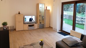 A modern living room with a flat-screen TV, wooden cabinets, a glass coffee table, and a view of the outdoors through a sliding glass door. - Ground floor apartment with large balcony. Terrace near the forest, to feel good (Schöneck)