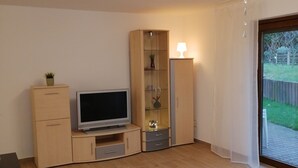 A modern living room with a flat-screen TV, wooden cabinets, a glass coffee table, and a view of the outdoors through a large window. - Ground floor apartment with large balcony. Terrace near the forest, to feel good (Schöneck)
