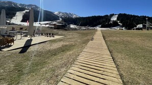 A clear sky with a sun shining, a wooden pathway leading to a seating area with tables and chairs, and a mountainous backdrop. - Studio Buron des Gardes (Le Lioran)