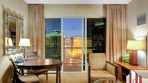 A hotel room with a dining table, two chairs, a sofa, a glass coffee table, a lamp, a telephone, and a view of the cityscape through a large window. - MGM Signature-29-607 F1 Track & Strip View Balcony (Las Vegas)
