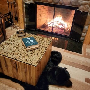 A cozy room with a fireplace, a wooden coffee table with a book, and a black dog lying on the floor. - The NEW Damon Pond Log Cabin (Amherst)