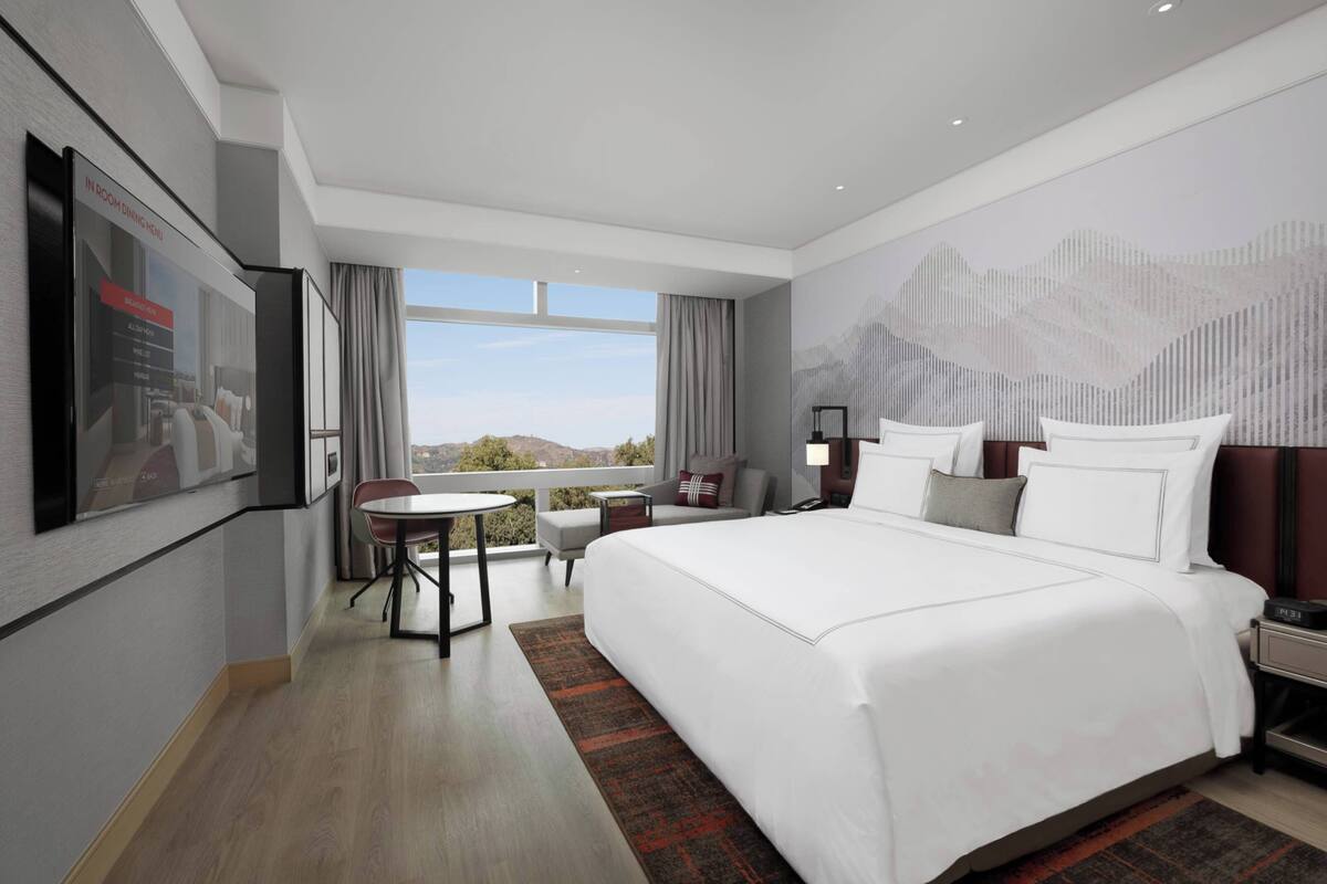 A modern hotel room with a large bed, a desk, a chair, and a view of the outdoors.