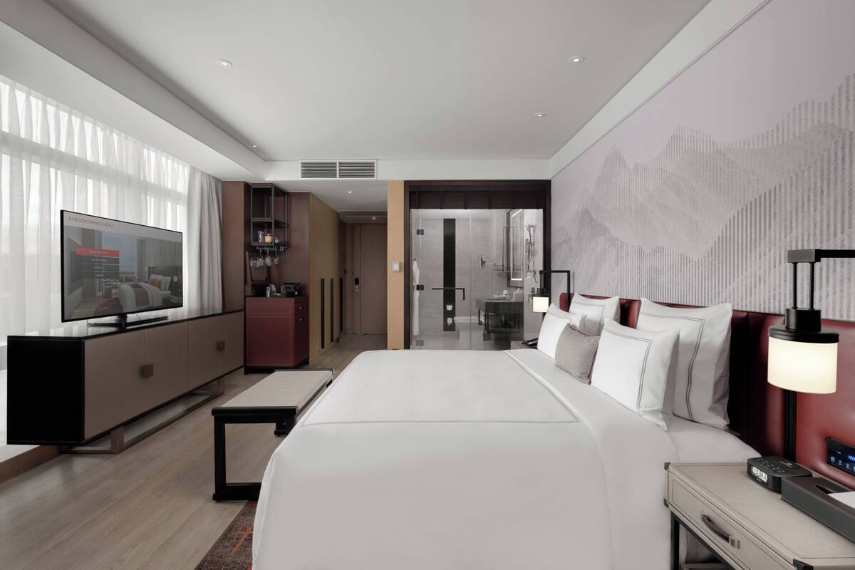 A modern hotel room with a large bed, a TV, a desk, and a bathroom visible through an open door.