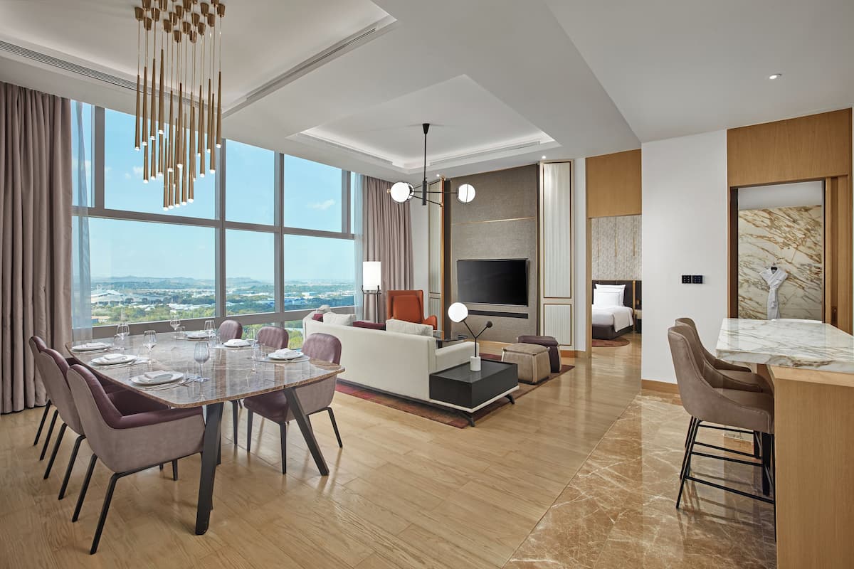 A modern hotel room with a dining area, a living room with a TV, and a large window offering a city view.