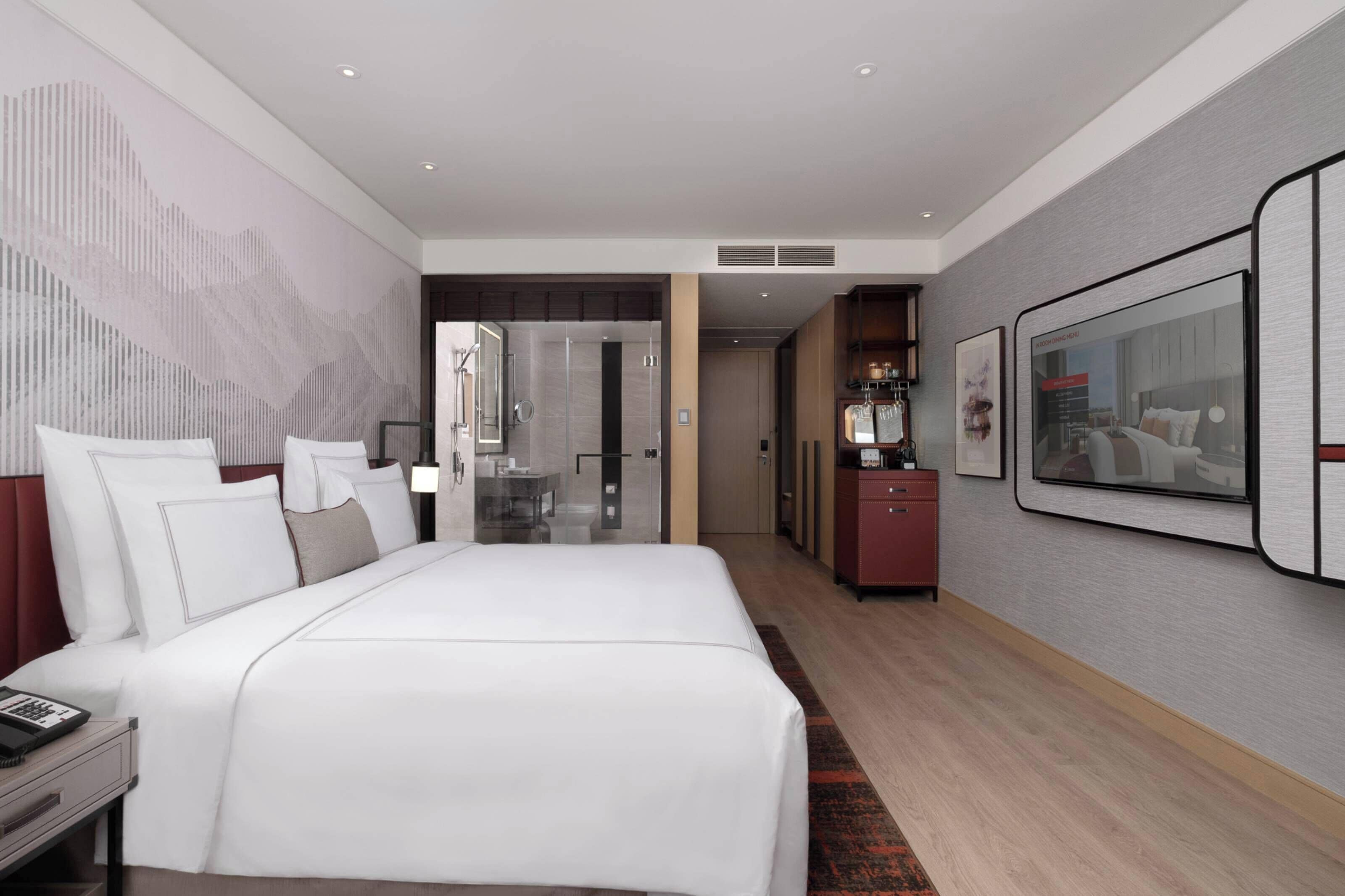 Une chambre d’hôtel moderne dotée d’un grand lit, d’un bureau, d’une armoire et d’une salle de bain visible à travers une porte en verre.