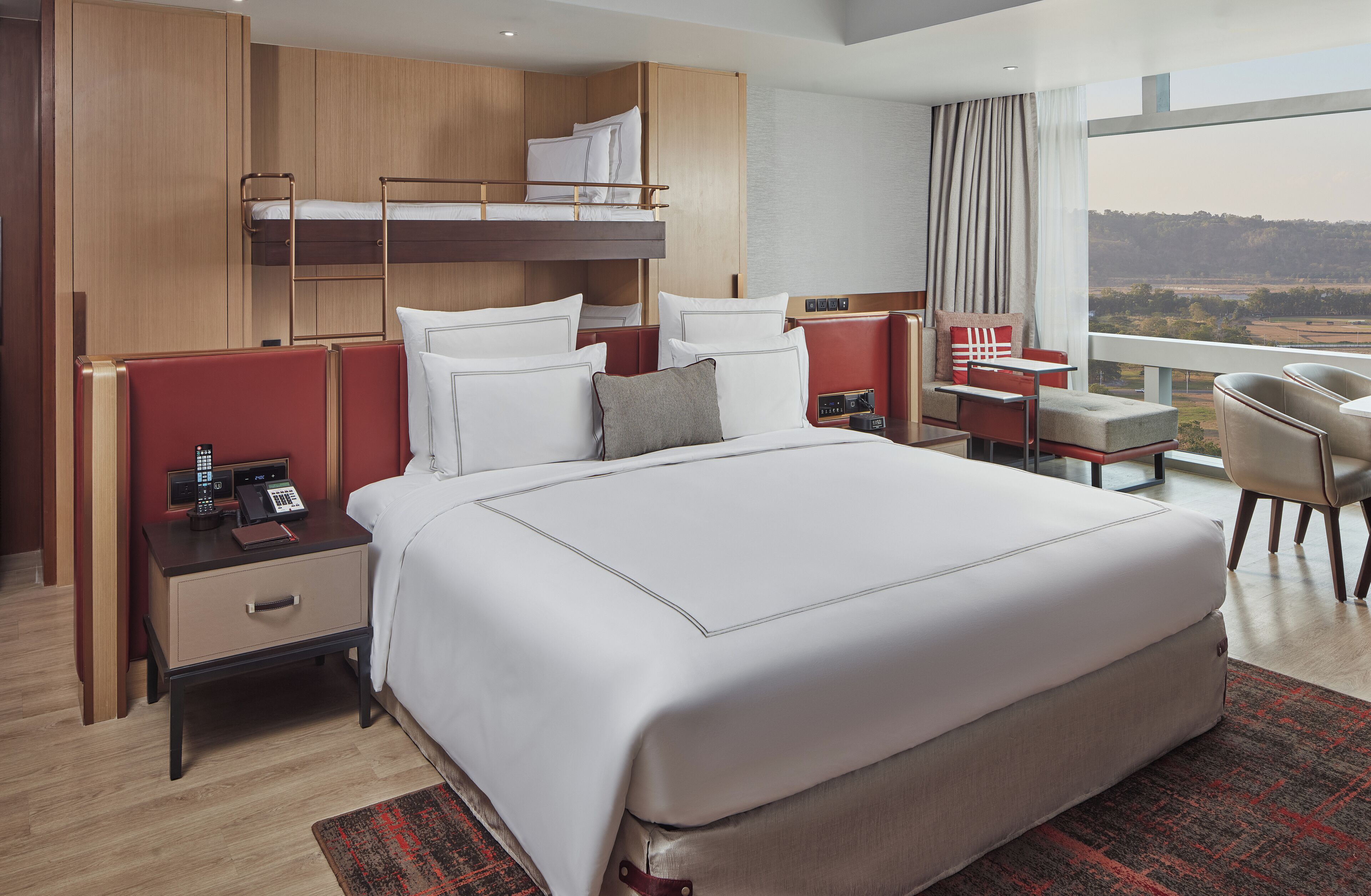 A modern hotel room with a large bed, a desk, a chair, a bunk bed, and a view of the landscape.