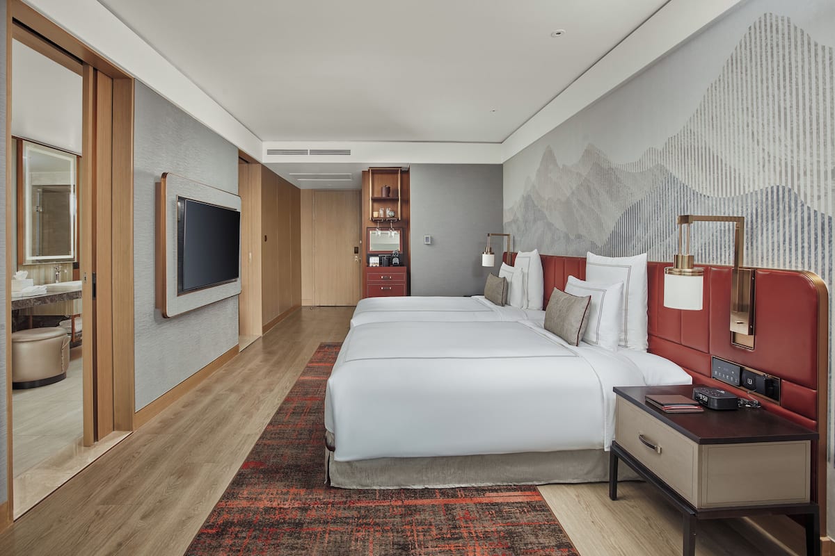 A hotel room with a large bed, a TV, a desk with a lamp, and a wall-mounted shelf.