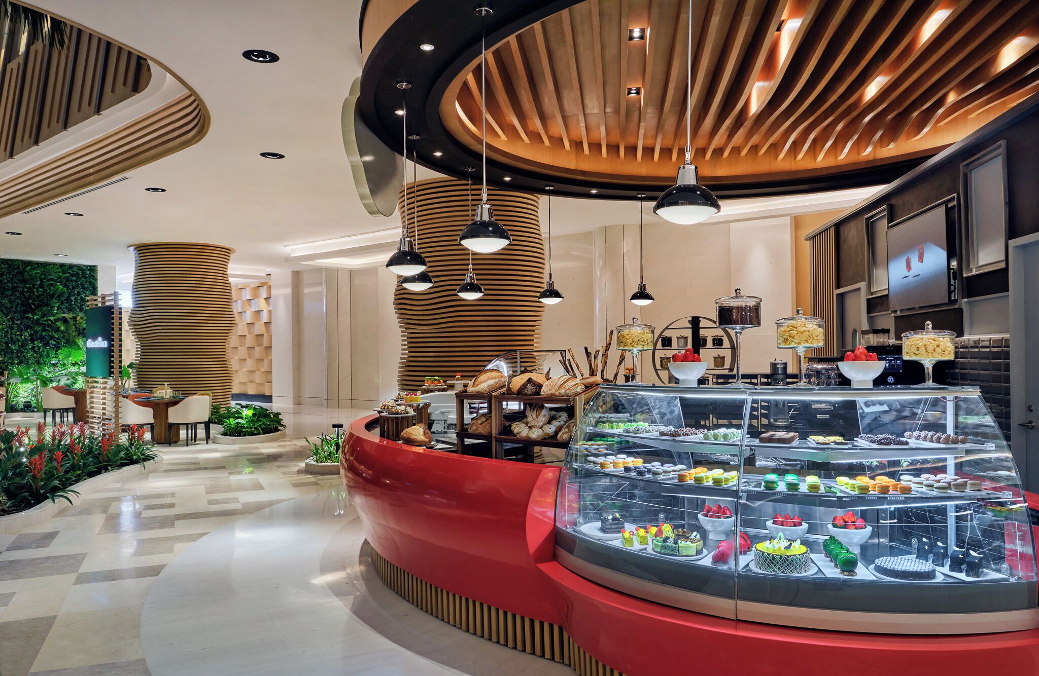 A modern hotel lobby with a central red seating area, a glass display case with pastries, and a wooden ceiling design.