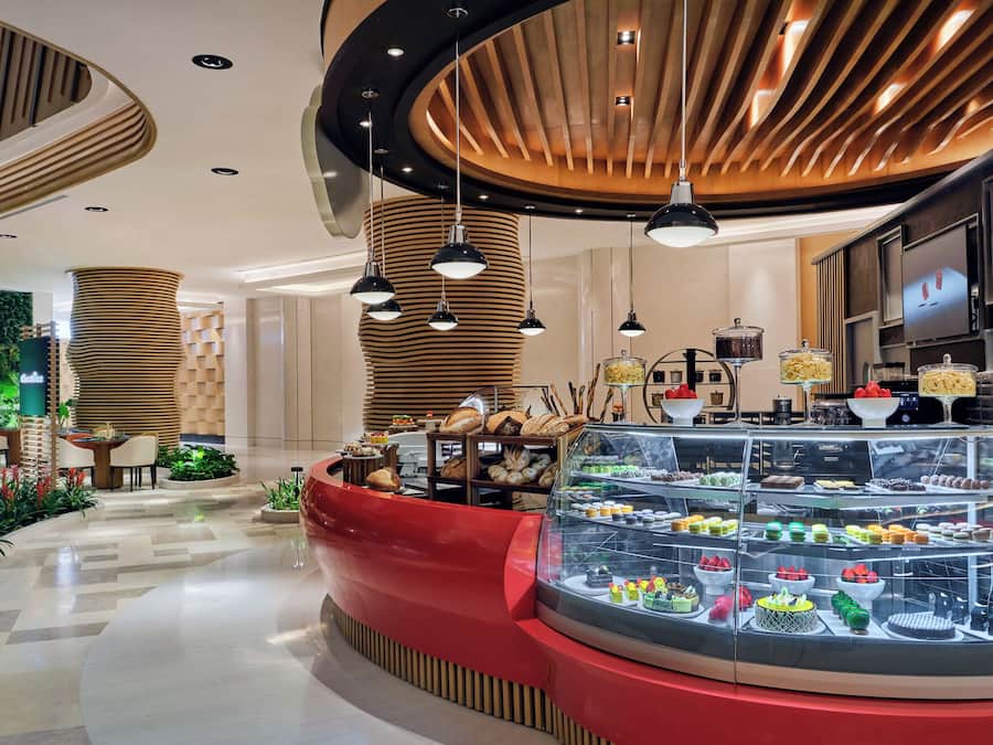 A modern hotel lobby with a central red seating area, a glass display case with pastries, and a wooden ceiling design.