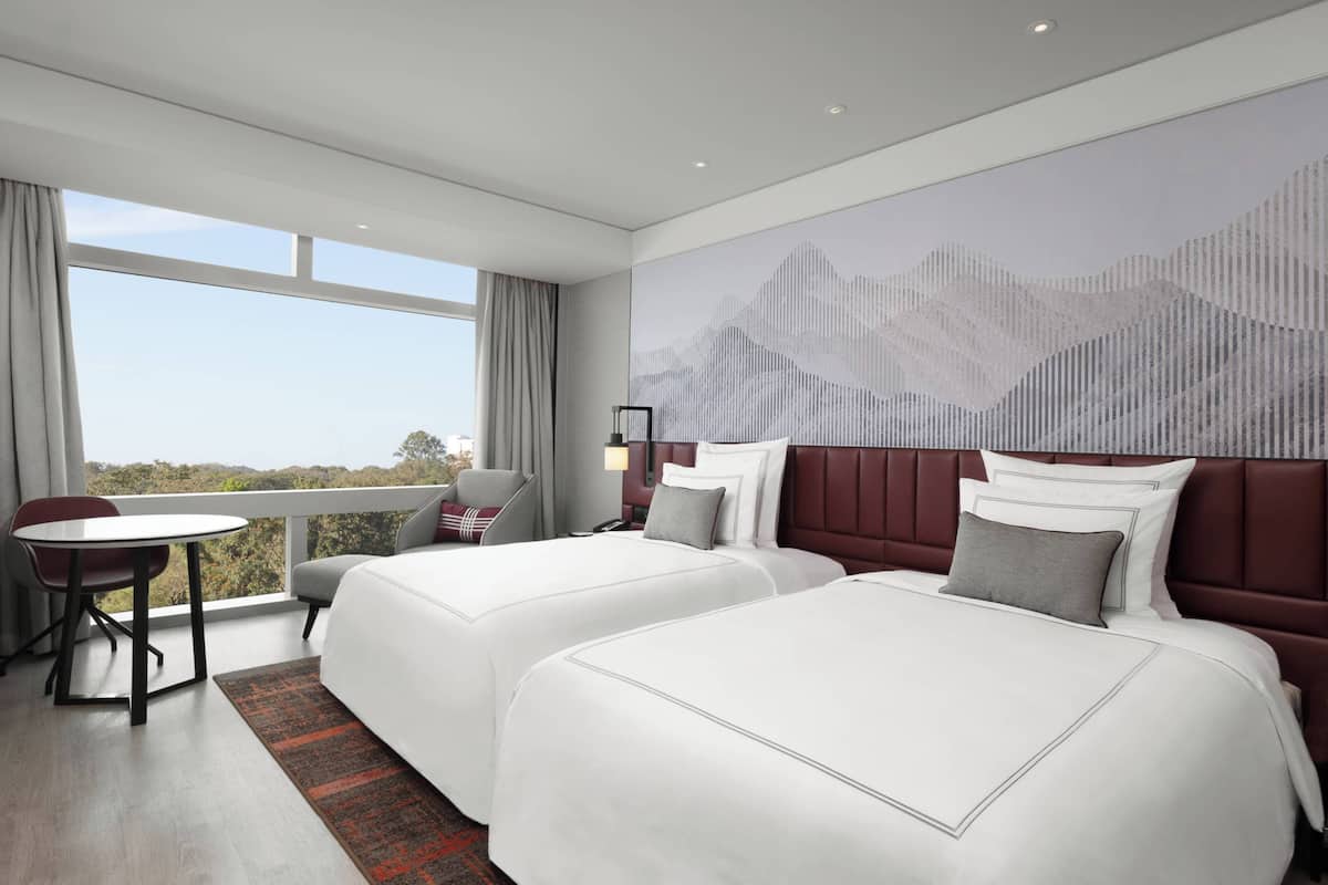 A hotel room with two beds, a desk, and a chair. There is a large window with a view of the outdoors.