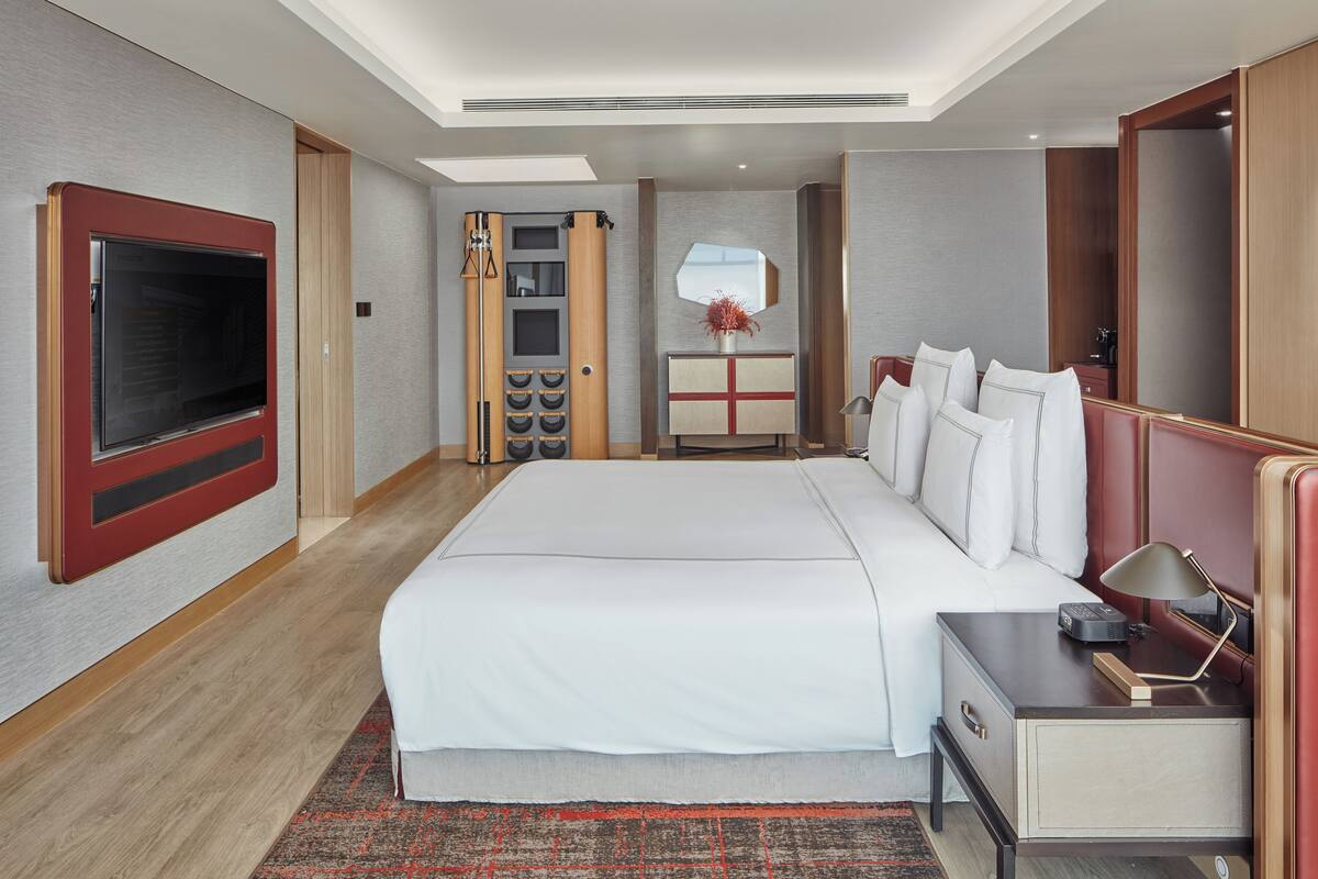 A modern hotel room with a large bed, a flat-screen TV mounted on the wall, a bedside table with a lamp, and a small desk.