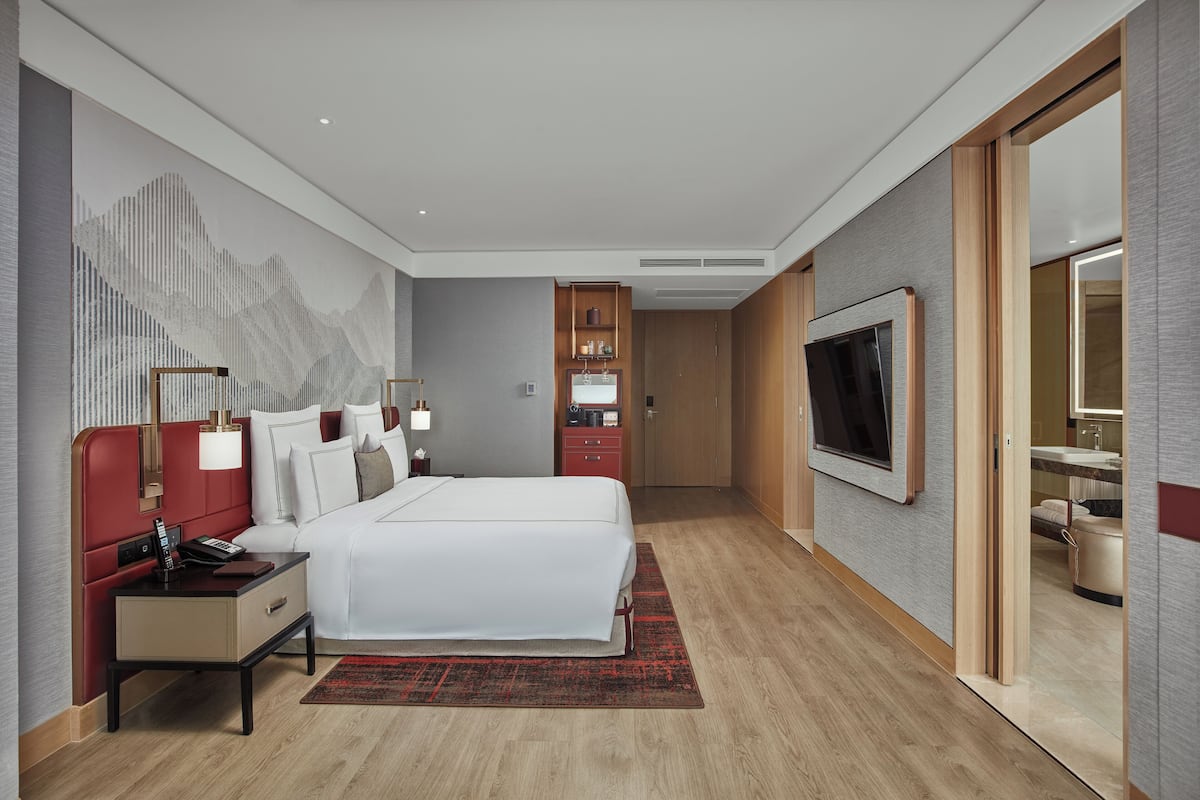 A modern hotel room with a large bed, a TV, a desk with a lamp, and a bathroom visible through an open door.