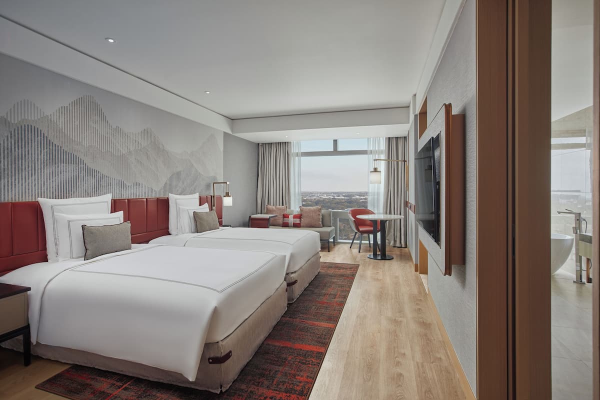 A modern hotel room with two beds, a TV, a desk, and a view of the city.