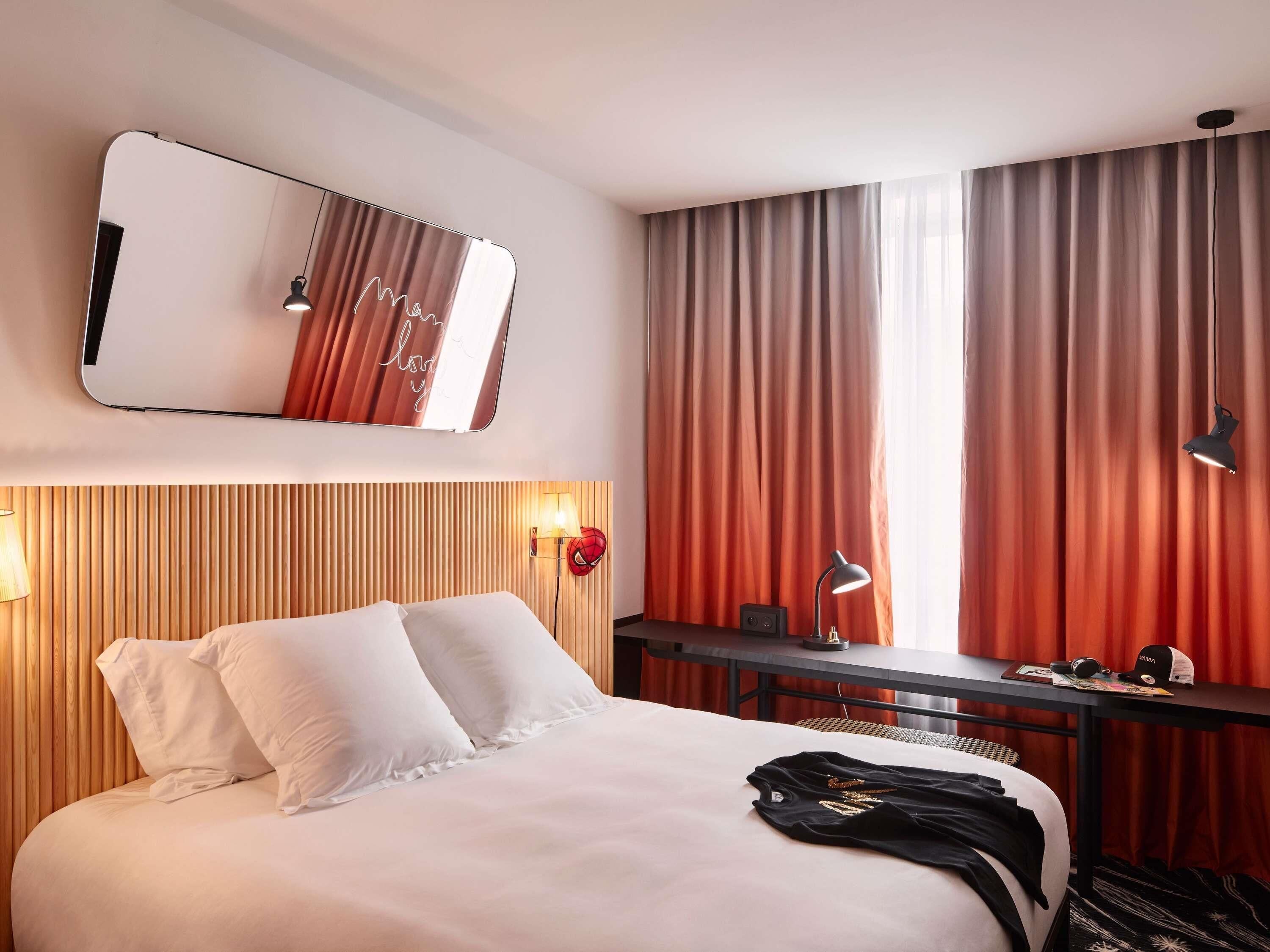 Une chambre d’hôtel avec un lit, un bureau, un miroir et une fenêtre avec des rideaux.