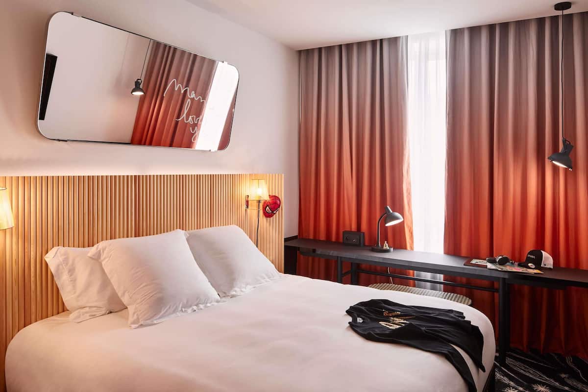A hotel room with a bed, a desk, a mirror, and a window with curtains.