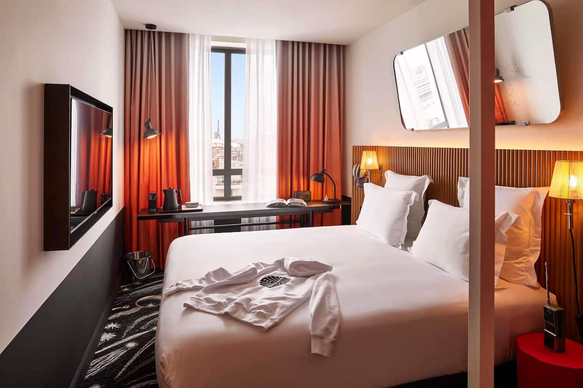 A hotel room with a bed, a desk with a chair, a large window with curtains, a mirror, and a wall-mounted TV.