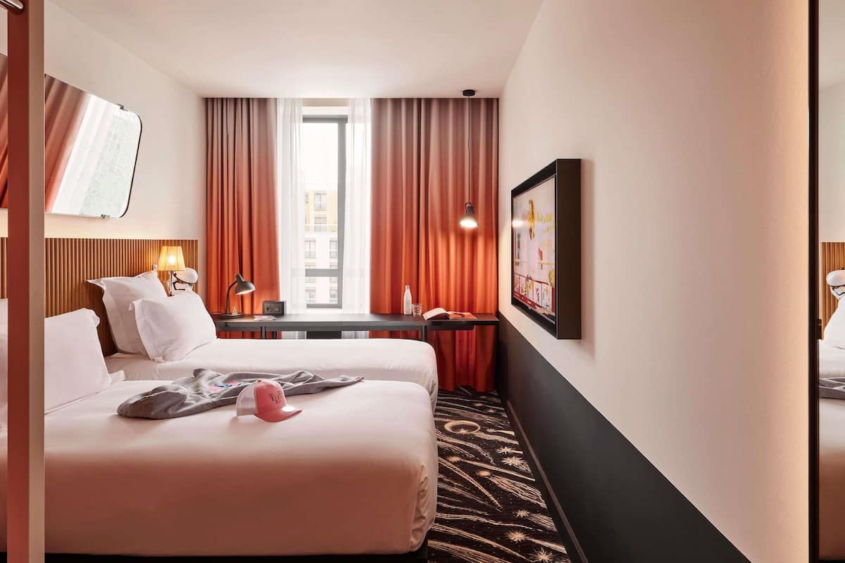 A hotel room with two beds, a desk, a lamp, a window with curtains, and a framed picture on the wall.