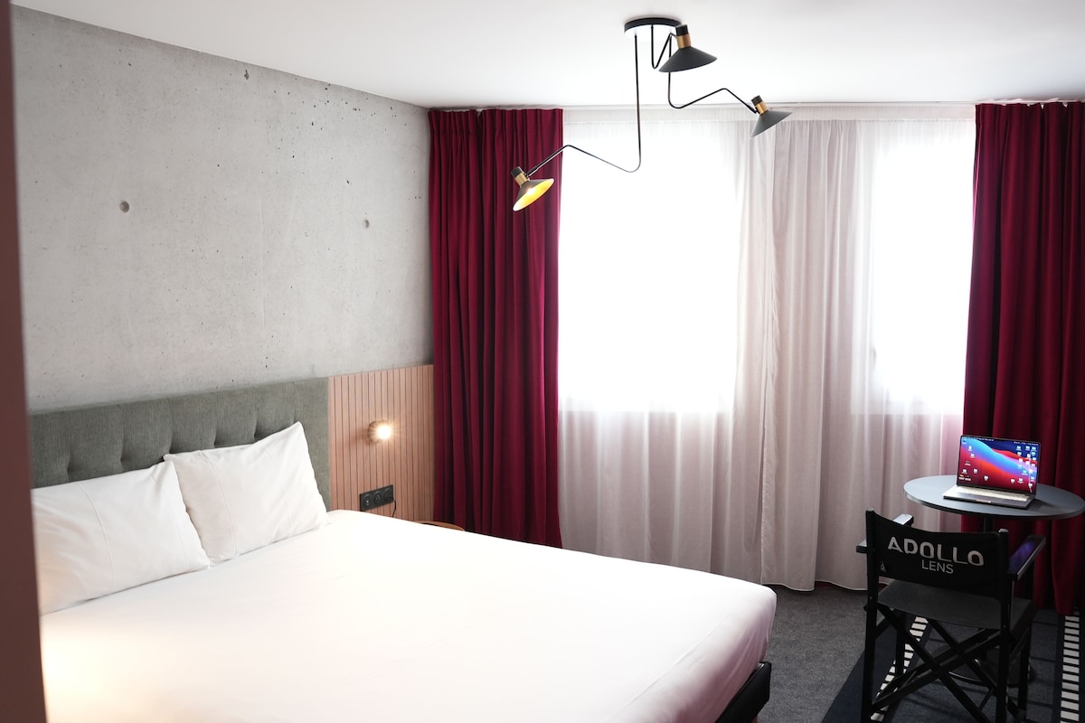 A hotel room with a bed, a desk with a laptop, a chair, and a lamp.