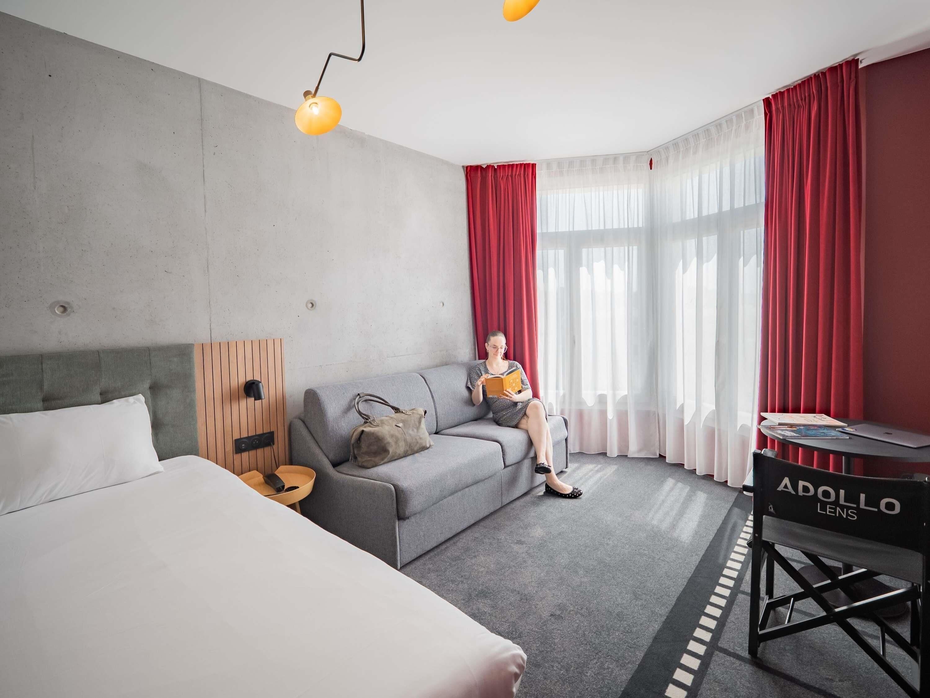 A hotel room with a grey sofa, a bed, a desk with a laptop, and a red curtain.