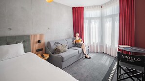A hotel room with a grey sofa, a bed, a desk with a laptop, and a red curtain. - Ibis Styles Lens Centre Gare (Lens)