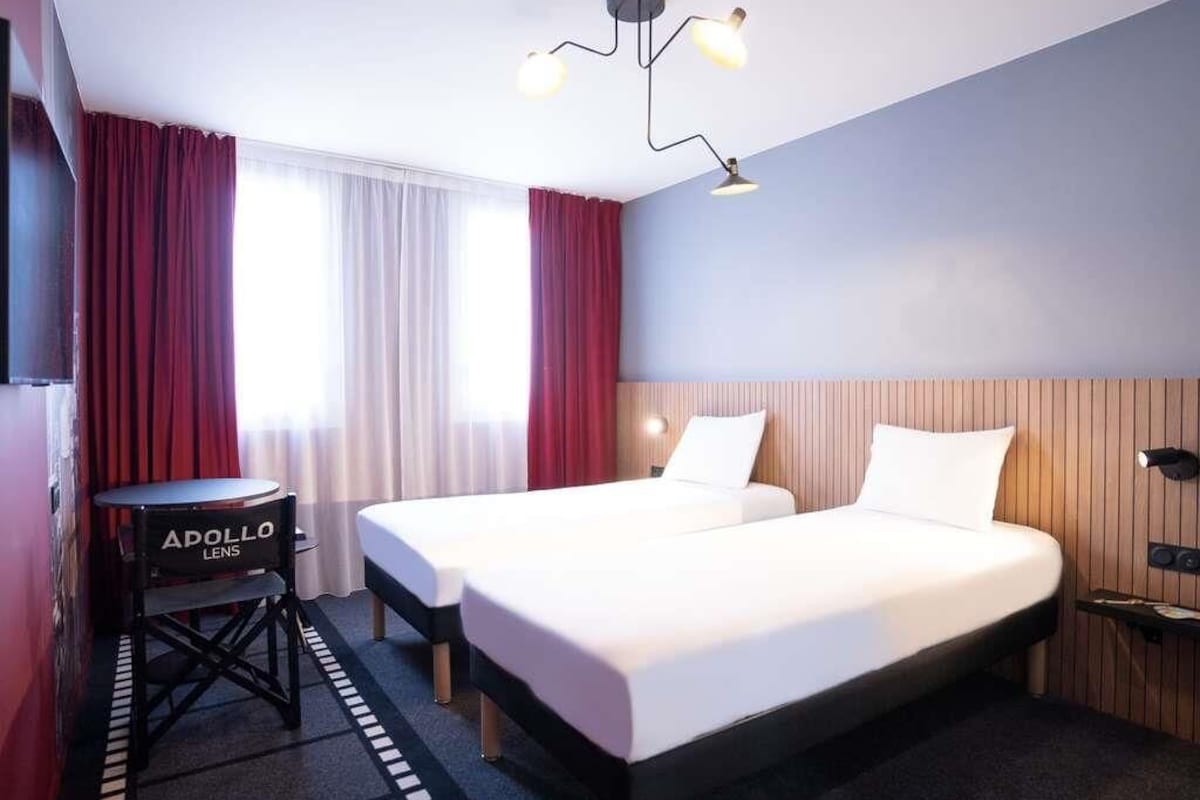 A hotel room with two beds, a desk with a chair, a television, and a modern chandelier.
