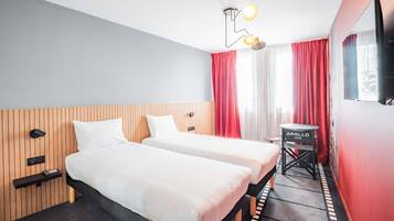 Habitación de hotel con dos camas, una mesita, un televisor y una ventana con cortinas rojas.